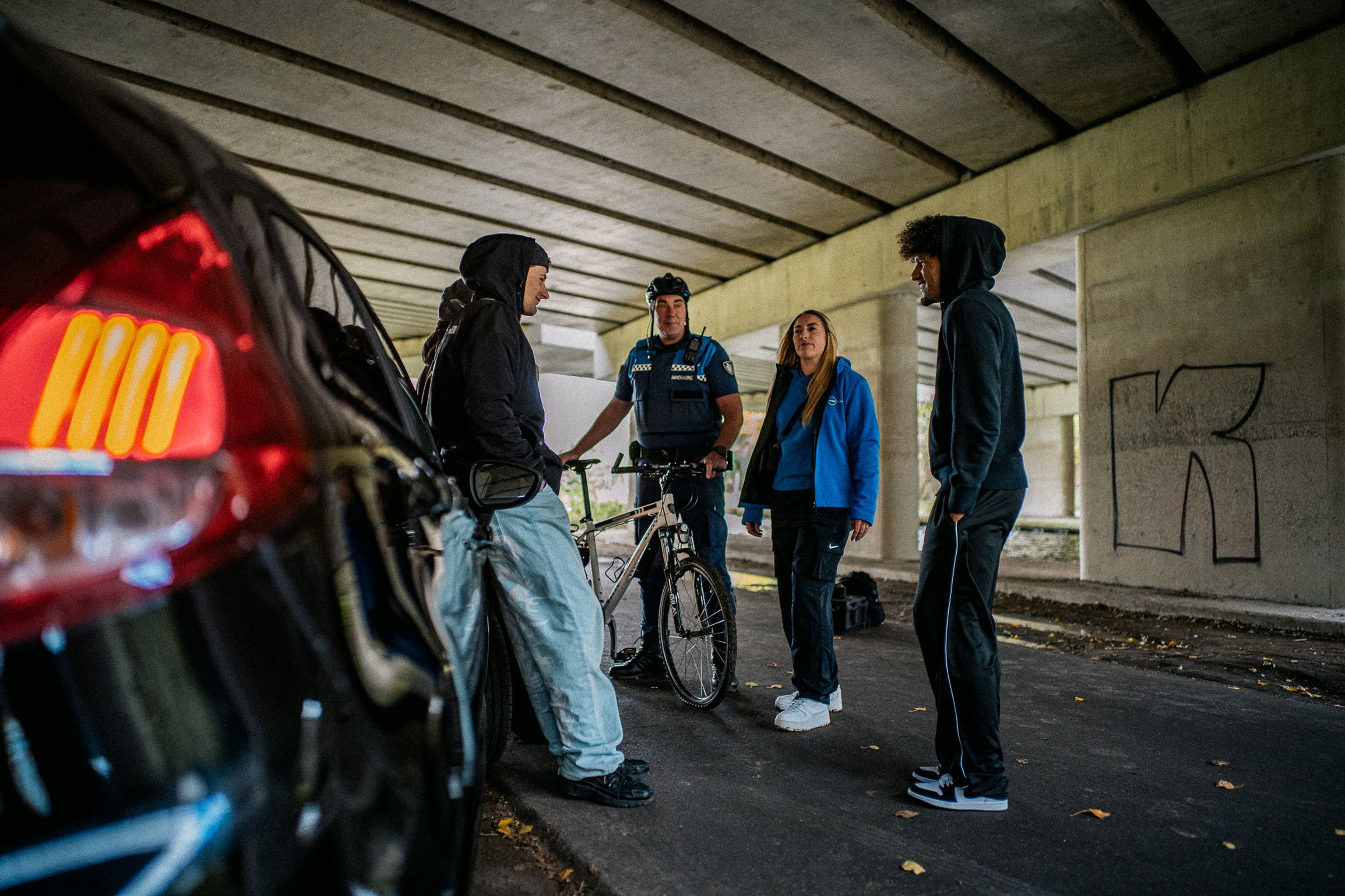 Jongerenwerker en Jeugdboa in gesprek met jongeren bij een auto onder een viaduct