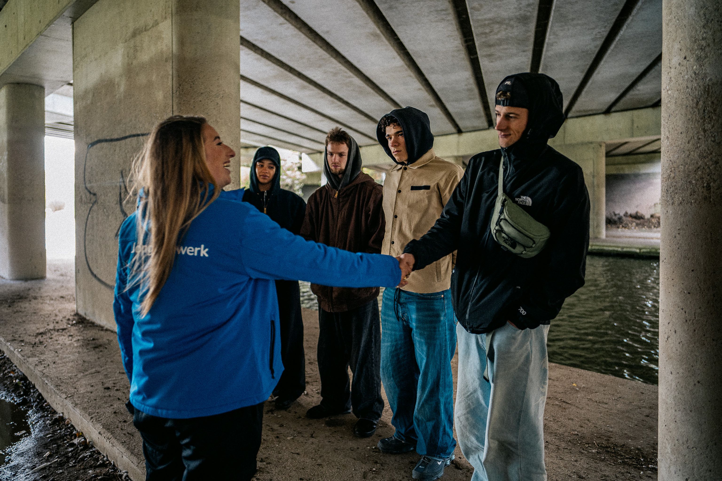 Groep jongeren in gesprek met jongerenwerker op een hangplek onder een viaduct. Jongerenwerker geeft hand aan jongen van jeugdgroep