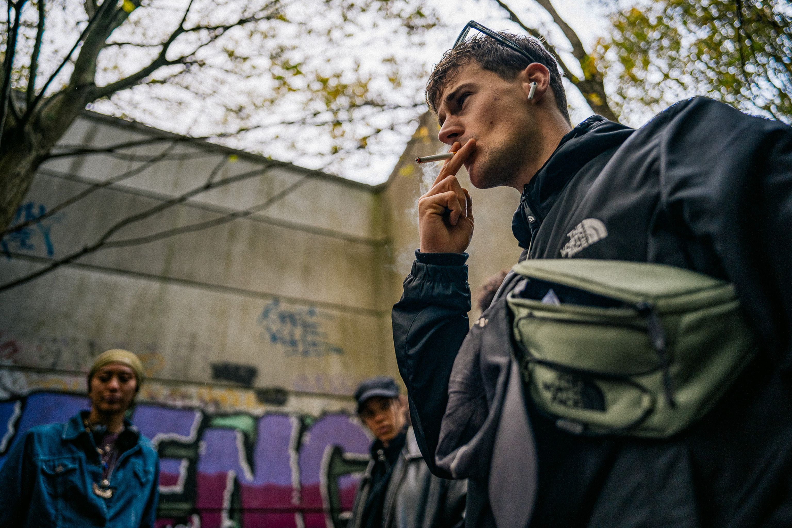 Jongere staat buiten met twee anderen op de achtergrond; hij houdt een sigaret vast terwijl de groep samen op een plek met graffiti rondhangt.